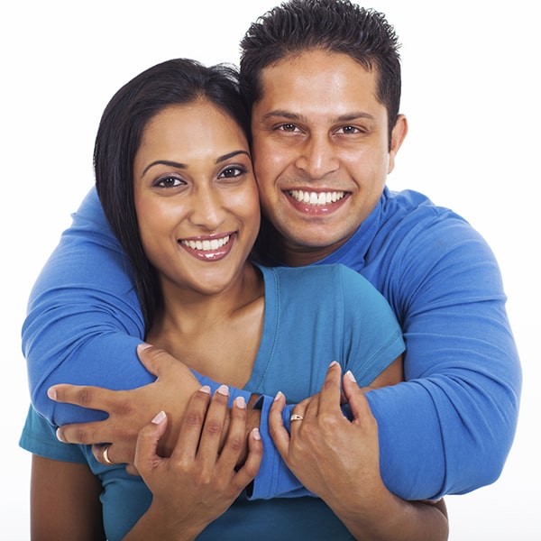 A smiling man back hugs a smiling woman while the woman holds on to the arms of the man.