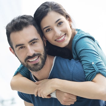 A smiling woman is back hugging a bearded smiling man