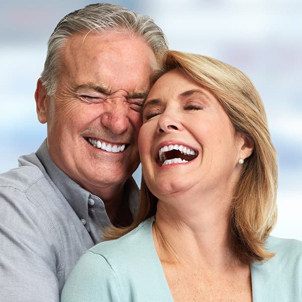 A smiling man back hugs a smiling  woman and leans her head back to the face of the man