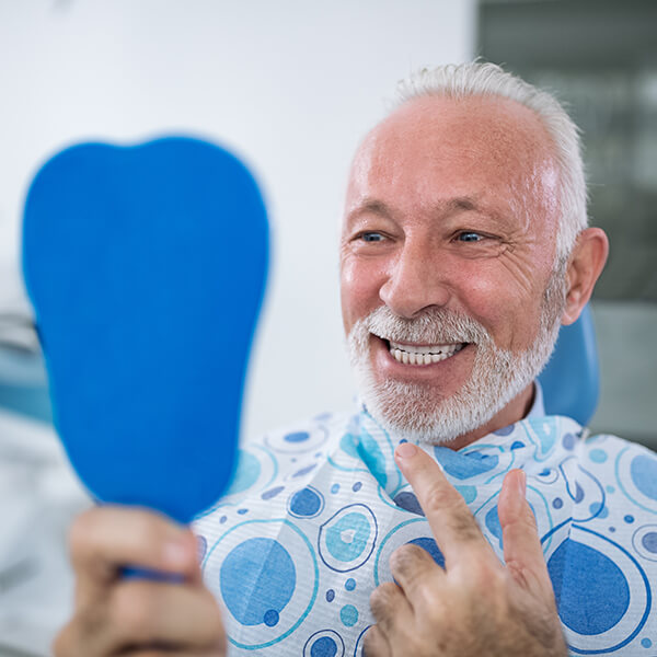 Senior man smiles as he looks into a hand mirror because he likes how his teeth look with restorative dentistry.
