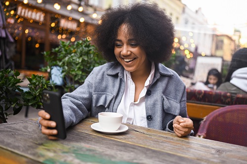 A young woman with straight teeth grins at her smartphone as she sits at an outdoor cafe.