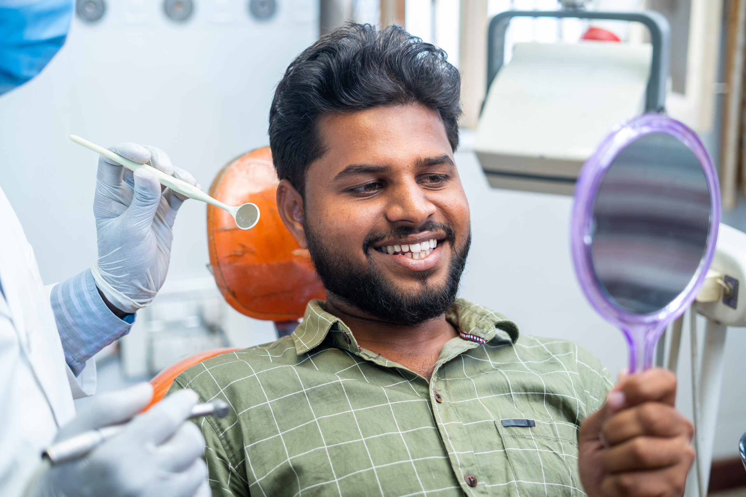 Young man with gleaming white teeth looks at his smile in a hand mirror after a teeth cleaning.
